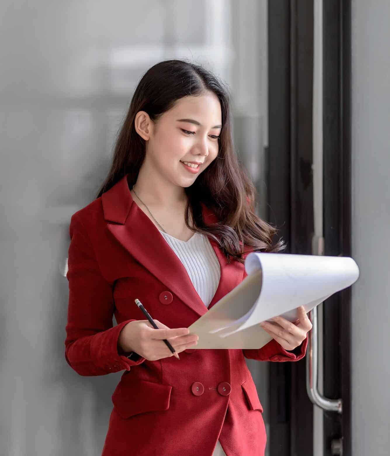 Happy young Asian businesswoman standing in a red suit holding and opening document in office.